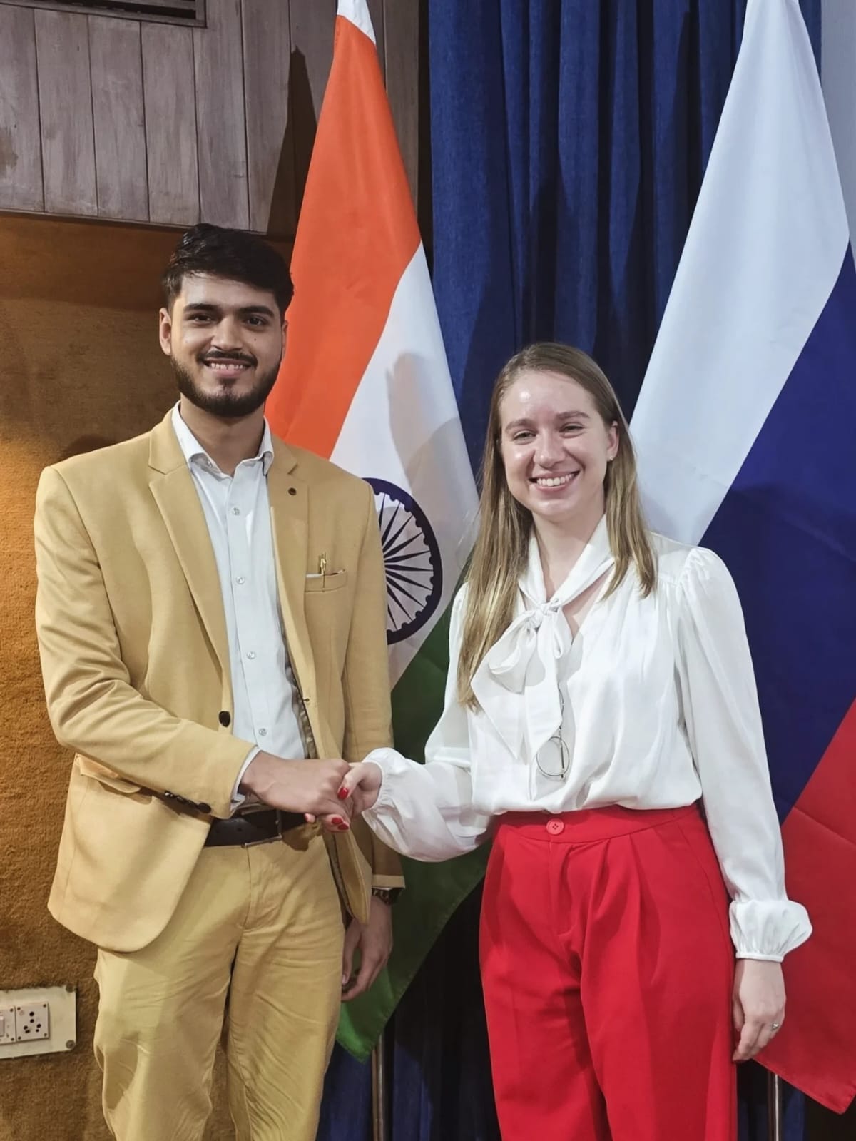 Akshat Mehrotra shaking hands with a female dignitary in front of flags.